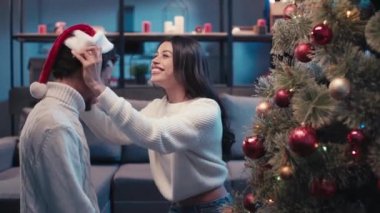 Afrika kökenli Amerikalı bir kadın Noel ağacının yanındaki sevgilisine Noel Baba şapkası takıyor.