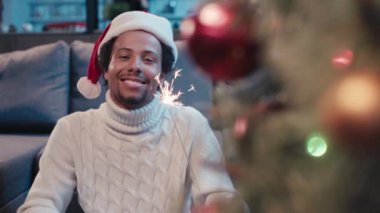 Noel Baba şapkalı mutlu Afro-Amerikan adam el sallıyor ve maytap tutuyor.