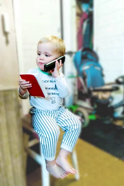 携帯電話の電話帳との子 アパート ミンスク市 2018 歳以下の子供は つの手で自宅の電話 もう一方の手でノートを保持します 彼は廊下の椅子に座っています ロイヤリティフリーのストック写真