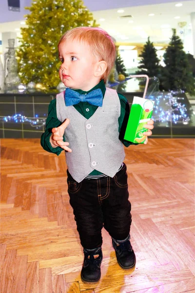 A boy at a Christmas party. Minsk city. 12/23/2018 He is wearing festive clothing: a green shirt with a butterfly tie, a gray vest, black pants and boots. He holds a bag of juice in his hand. Behind him is an elegant Christmas tree with a garland.