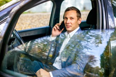 Yakışıklı adam akıllı telefon tutarak ve gülümseyen arabada oturan takım elbiseli.