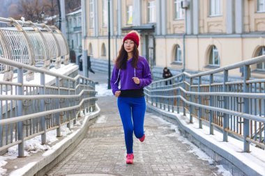 Bir şapka ve eşofman atletik Asyalı kız şehirde bir köprü üzerinde çalışır. Sağlıklı yaşam ve spor. Güzel kız kış aylarında egzersizleri yapıyor.