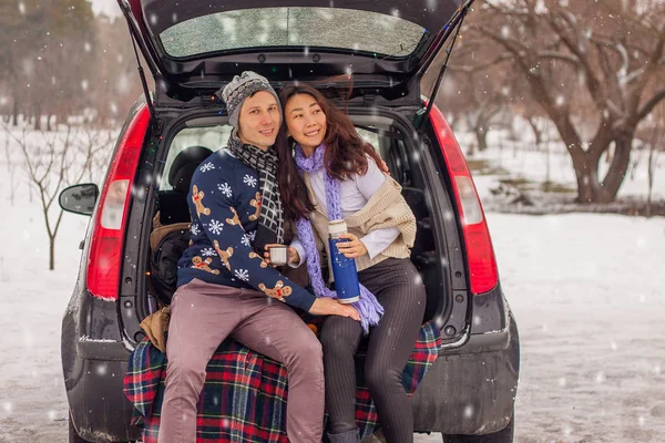 Beautiful international couple relaxing in nature in winter. Romantic meeting of a couple in love in the snow. Young couple sit in the trunk and drink tea.