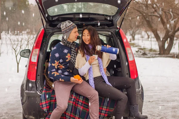 Beautiful international couple relaxing in nature in winter. Romantic meeting of a couple in love in the snow. Young couple sit in the trunk and drink tea.