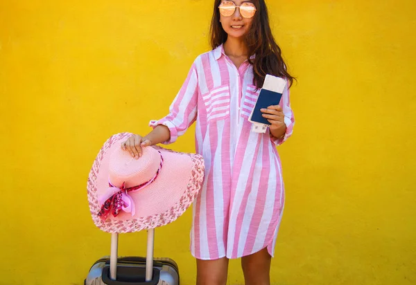 Happy Asian woman with a suitcase and a ticket is standing near the yellow wall.