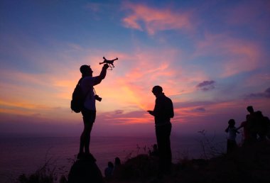 Silhouette of traveler controlling copter and photographing colorful sunrise. Man makes aerial photo and video of sunset sky on beach. Tourist holds remote control fpv. clipart