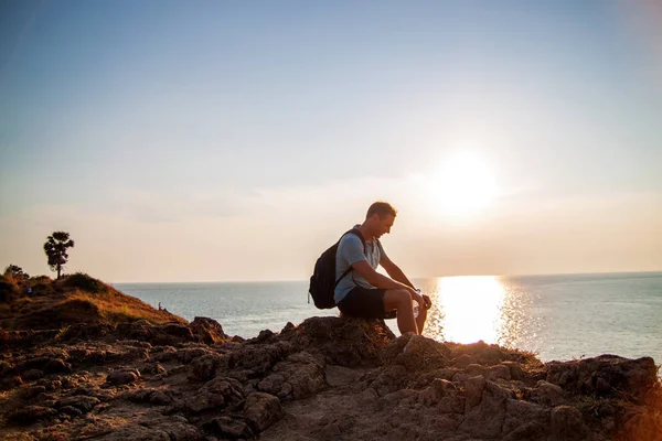 Side view of man with backpack looking at sea while sitting on hill against sky during sunny day.