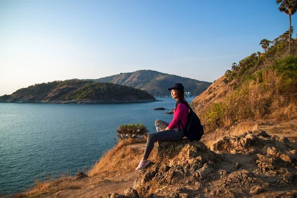 Side view of a happy woman with backpack looking at sea while sitting on hill against sky during sunny day.