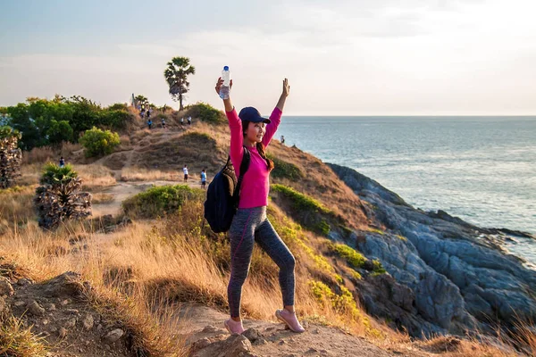 Tourist with backpack and a bottle of water standing on top of a hill with raised hands at sunset.