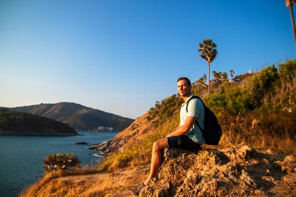 Side view of a happy man with backpack looking at sea while sitting on hill against sky during sunny day.