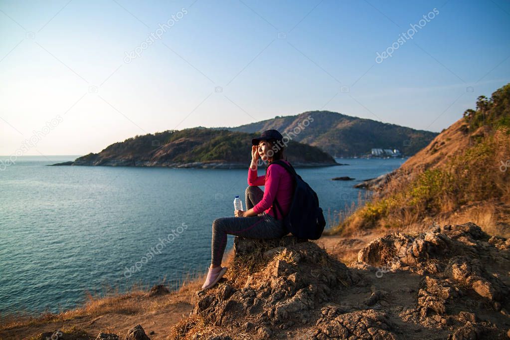 Side view of a happy woman with backpack looking at sea while sitting on hill against sky during sunny day.