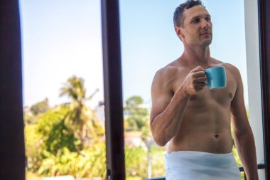 Yakışıklı gömleksiz kaslı genç adam balkonda sabahın erken saatlerinde kahve içme ve görünümün tadını çıkarın. 