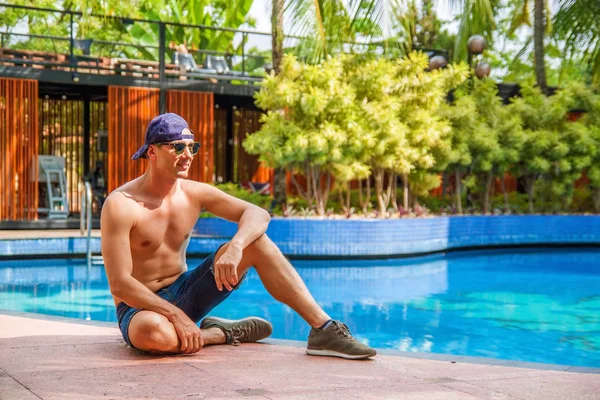 Yakışıklı Genç Adam Yüzme Havuzu kenarına eğilerek ve Tropikal İklim kamera gülümseyerek.
