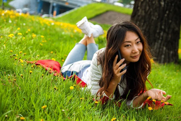 Asian happy woman calling by phone in summer park on green grass.