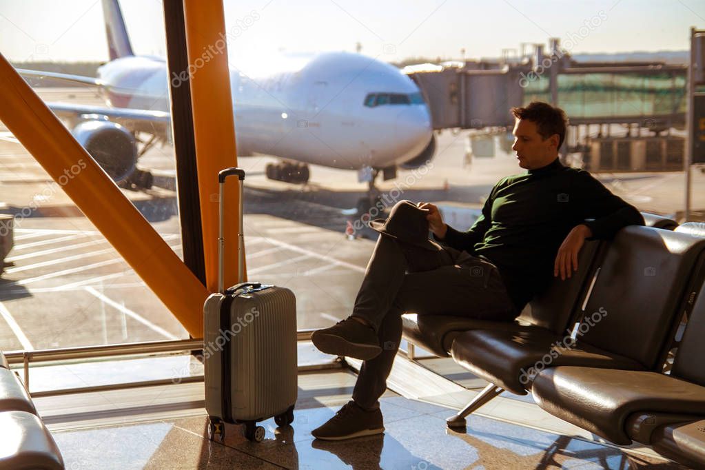 Handsome young man talking on the phone, sitting on a chair with things at the airport waiting for his flight.