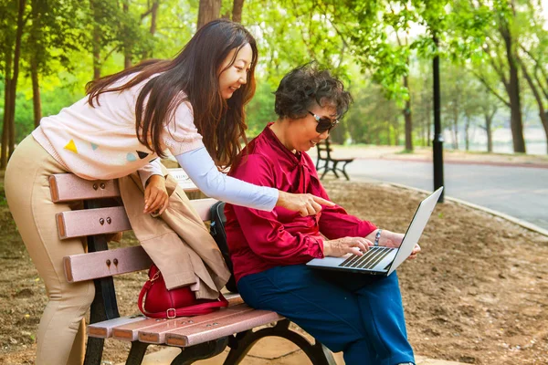 a smiling mom and daughter using a laptop at garden. Daughter teaches an elderly mother to use a computer.