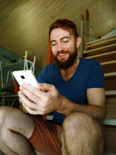 Portrait of a young handsome redhead man with a beard relaxing at home using by mobile phone with a big smile .