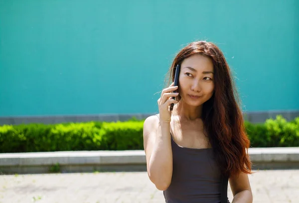 Asian cute girl talking over mobile phone while waking in the street. Cheerful young girl talking over cell phone standing outdoors in a park.