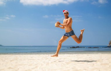 Tatilde noel baba şapkası takarak sahilde zıplayan yakışıklı genç adam.