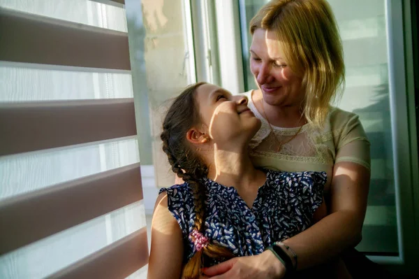 Mother and daughter sitting on sill near window in room. They show emotions and have fun.