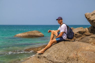 Deniz kenarında sırt çantası olan atletik adam arkasında deniz olan birini aramak için cep telefonu kullanarak, bir kayanın üzerinde oturan