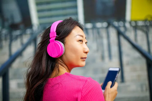 Waist up portrait of happy young asian woman training. She is listening to music from earphones and looking at smartphone. Athlete is standing outdoors and smiling.