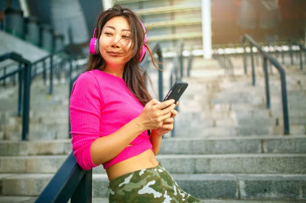 Waist up portrait of happy young asian woman training. She is listening to music from earphones and looking at smartphone. Athlete is standing outdoors and smiling.
