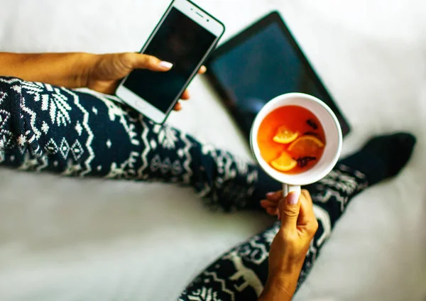 Girl sitting on furry carpet in home clothes and Christmas socks. Woman holds cup of tea on leg and smartphone.  Christmas and New Year concept.