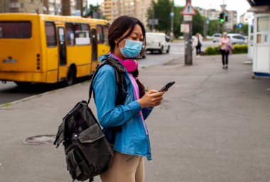 Otobüs durağında otobüs bekleyen Çinli bir kadın var. Yüzünde maske var.