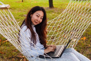 Genç Asyalı Kadın Sonbahar Parkı 'nda Hamakta Dizüstü Bilgisayar Kullanıyor.