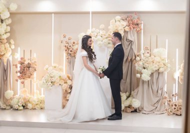 Düğün manzarası, takım elbiseli bir adam çiçek kemerinin yanındaki beyaz kabarık elbiseli bir kadını öpüyor..