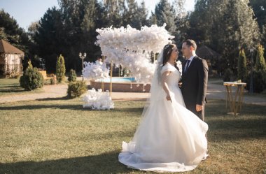 Takım elbiseli şık bir damat ve çiçeklerle süslenmiş bir düğün kemerinin yanındaki ormanda beyaz elbiseli tatlı esmer bir gelin. Yeni evlilerin düğün portresi..