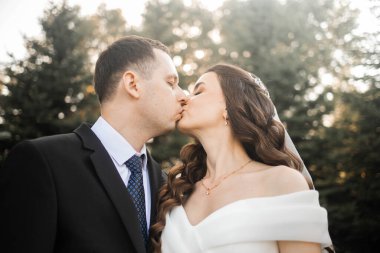 Düğün çifti gelin ve damat çam ormanlarında sarılıyorlar, güneş ışığı ağaçların arasından süzülüyor, neşeli ifadelerini vurguluyorlar..