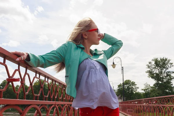 Young blond woman in leather jacket and glasses posing on red bridge looking into the sky. The wind blows the hair