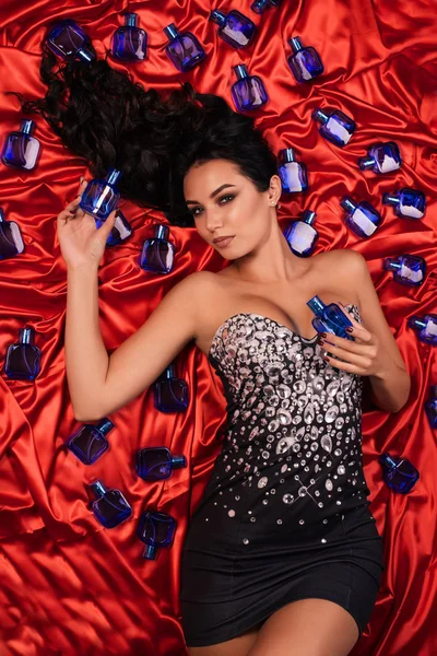 Beautiful young woman in a shiny dress posing on red satin surrounded by perfume bottles.