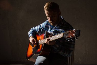 Ekose gömlekli sakallı bir adam akustik gitar çalar.