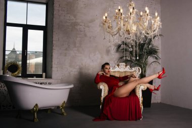 Güzel kadın oturmuş bir sandalyede Her bacaklar yukarı ve bir Vintage Red ilham bohem bir pencere ve küvet yanındaki elbise ile