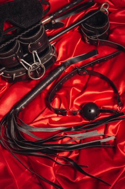 Bondage, kinky adult sex games, kink and BDSM lifestyle concept with a pair of leather handcuffs, flogger, ball gag and a coller with a leash attached on red silk with copy space clipart