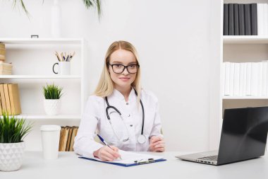 Genç, güzel doktor kadın mutlu çalışıyor ve hastanede gülümsüyor, masada oturuyor.