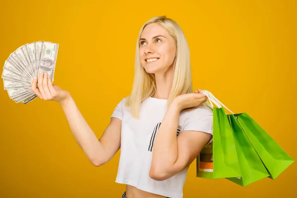 Närbild porträtt av lycklig ung vacker blond kvinna som innehar pengar och gröna shopping väskor, isolerade på gul bakgrund — Stockfoto