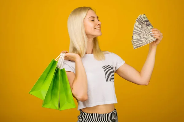 Närbild porträtt av lycklig ung vacker blond kvinna som innehar pengar och gröna shopping väskor, isolerade på gul bakgrund — Stockfoto