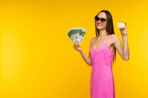 Menina espanhola magra em um vestido rosa e óculos de sol segurando uma centena de notas de euro e um cartão de crédito — Fotografia de Stock