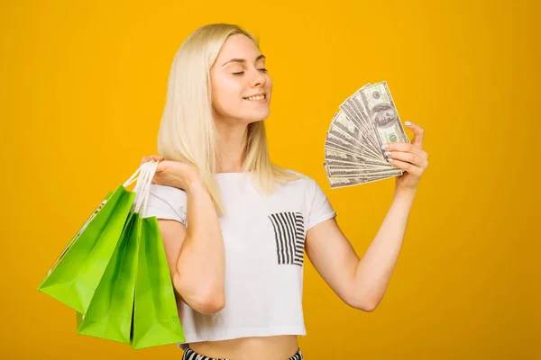 Närbild porträtt av lycklig ung vacker blond kvinna som innehar pengar och gröna shopping väskor, isolerade på gul bakgrund — Stockfoto