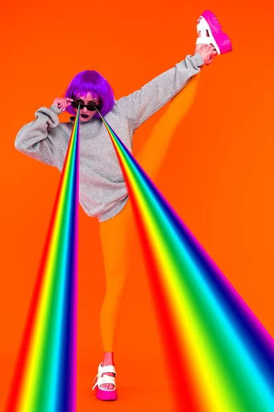 Menina lésbica em uma peruca roxa e óculos de sol dispara lasers de seus olhos arco-íris em pé sobre um fundo laranja tendo levantado uma perna para cima . — Fotografia de Stock