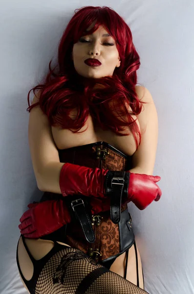 Κοκκινομάλλα γυναίκα σε μια κόκκινη περούκα, κορσέ και δερμάτινα γάντια που θέτουν σαγηνευτικά κείτεται στο κρεβάτι — Φωτογραφία Αρχείου