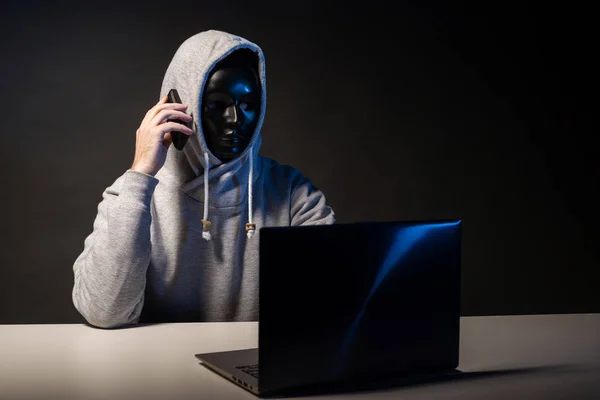 Анонимный хакер в маске программист использует ноутбук и разговаривает по телефону, чтобы взломать систему в темноте . — стоковое фото