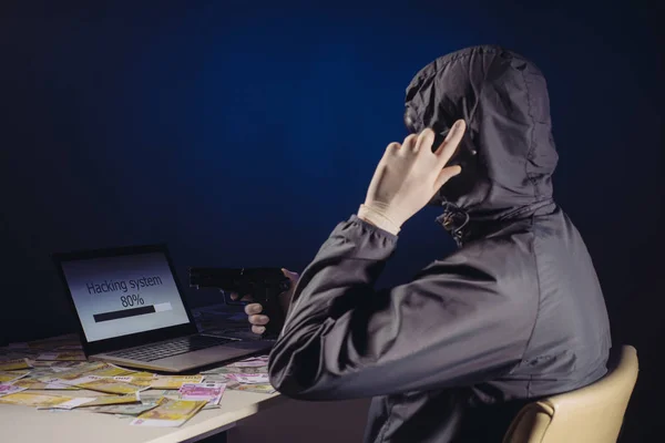Programador de hackers anônimo usa um laptop segurando uma arma em sua mão para invadir o sistema no escuro. Criação e infecção de vírus malicioso . — Fotografia de Stock