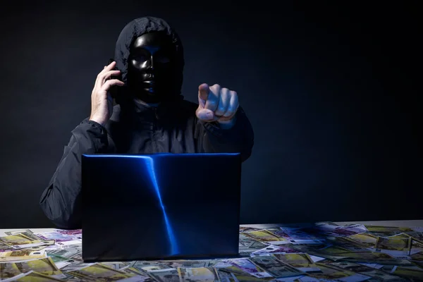 Hacker anônimo no programador máscara usa um laptop e falando no telefone para hackear o sistema no escuro . — Fotografia de Stock