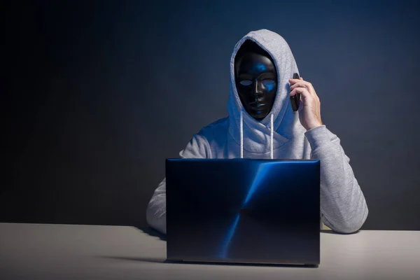 Anonymous hacker in mask programmer uses a laptop and talking on the phone to hack the system in the dark.