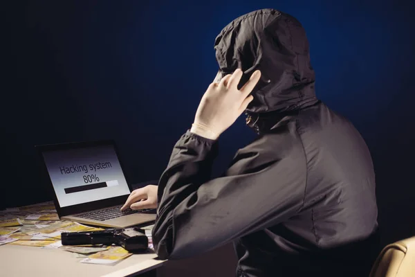 Hacker anônimo na máscara sentado lateralmente usa um laptop e falando no telefone para cortar o sistema no escuro. O conceito de cibercrime e base de dados de pirataria — Fotografia de Stock